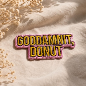 Puede incluir: Un parche bordado que presenta el texto "GODDAMNIT, DONUT" en letras amarillas en negrita con un contorno negro. Las letras están sobre un fondo rosa con un borde festoneado, creando una forma rectangular distintiva de dos líneas.