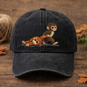 Peut inclure: Casquette de baseball en denim noir avec un motif brodé de deux singes de dessin animé. L'un tire l'autre, qui est allongé. La casquette a une visière incurvée et un bouton sur le dessus.