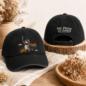 Può includere: Cappellino da baseball nero con un procione ricamato che indossa un mantello arcobaleno e tiene una spada. Il cappellino presenta la scritta "WE PRIDE AT DAWN" sul retro. Il design include un'alba e delle nuvole.