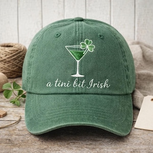 May include: Green baseball cap with an embroidered martini glass and shamrock design. The text "a tini bit Irish" is embroidered below the design. The cap is made of a soft, textured fabric.