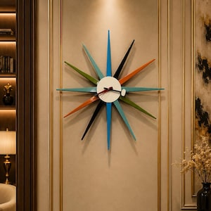 May include: A starburst wall clock with a white face and colorful, pointed arms in blue, green, orange, and black. The clock is mounted on a beige wall with gold trim, adding a mid-century modern touch to home decor.