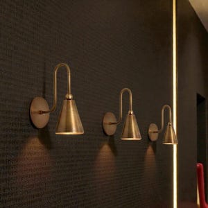 May include: Three brass wall sconces with a conical shade and curved arm are mounted on a dark textured wall. The sconces are of varying sizes, with the largest on the left. A vertical light strip is on the right.