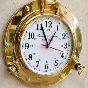 May include: A round wall clock with a polished brass porthole frame. The white clock face features black Arabic numerals from 1 to 12, smaller 13 to 24 hour markers, and the text "Franke & Murphy EST 1883". It has black hour and minute hands and a red second hand.