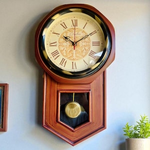 Op de afbeelding: Een vintage wandklok met een houten frame en een crèmekleurige wijzerplaat. De klok heeft Romeinse cijfers, decoratieve wijzers en een slinger. De klok heeft een donkerbruine rand en een rechthoekig onderste gedeelte met een glazen venster.