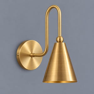 May include: A wall-mounted light fixture with a brushed brass finish. It features a circular wall plate, a curved arm that extends upward and then downward, and a conical shade that directs light downward. The metal has a smooth, metallic texture.