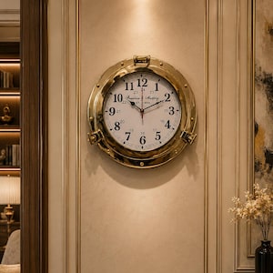 May include: A brass-colored wall clock with a white face and black numerals. The clock has a nautical porthole design, with the text "Franklin & Murphy" and "ST 1868" visible. The clock is mounted on a beige wall with gold trim.