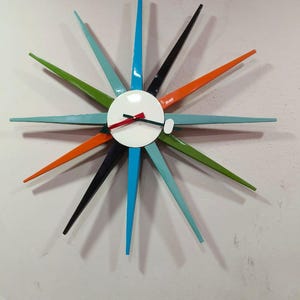 Puede incluir: Un reloj de pared retro con forma de estrella, con una esfera blanca y brazos puntiagudos de colores. Los brazos son en tonos de azul, verde, naranja y negro. Las manecillas del reloj son rojas y negras.