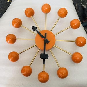 May include: A sunburst wall clock featuring a central orange circular face with twelve thin gold-toned metal spokes radiating outward. Each spoke is tipped with a smooth orange sphere. The clock has black hour and minute hands and a red second hand.