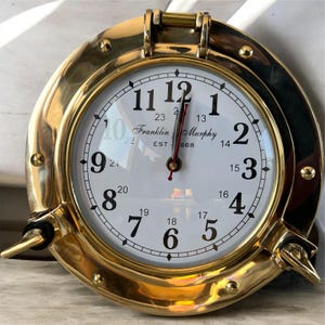 May include: A brass-colored, round clock with a white face and black numerals. The clock has a nautical porthole design with a hinged top and side latches. The clock face reads "Franklin Murphy EST 1868".
