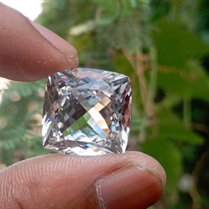 Può includere: Una pietra preziosa trasparente e incolore, con taglio a cuscino e una superficie sfaccettata che riflette la luce. La pietra di forma quadrata presenta un motivo a taglio brillante con numerose sfaccettature triangolari e a rombo.