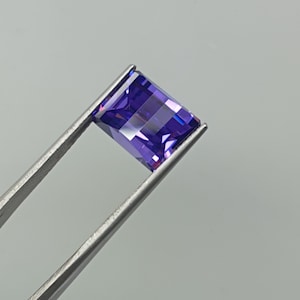 Pode incluir: Uma pedra preciosa retangular facetada com um tom roxo profundo, segurada por uma pinça de metal prateado. A pedra apresenta uma qualidade clara e transparente com bordas afiadas e uma superfície polida que reflete a luz.