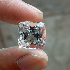 Può includere: Una pietra preziosa taglio cuscino con un aspetto chiaro e trasparente e una superficie sfaccettata che riflette la luce. La pietra presenta una forma quadrata con angoli arrotondati e una qualità brillante e riflettente in tutta la sua struttura.