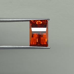 Puede incluir: Una piedra preciosa naranja rectangular y facetada sujeta por unas pinzas metálicas. La piedra presenta un tono naranja vibrante y profundo con una superficie clara y pulida, y bordes geométricos afilados que reflejan la luz en su estructura.