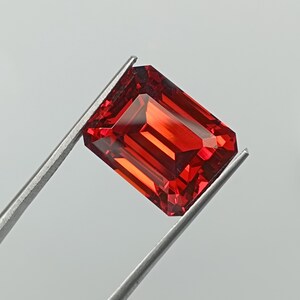 Pode incluir: Uma gema de cor vermelha profunda e vibrante com corte esmeralda, apresentando facetas nítidas e limpas que refletem a luz. A pedra retangular é mantida por uma pinça de metal prateado, exibindo a sua cor transparente e saturada.
