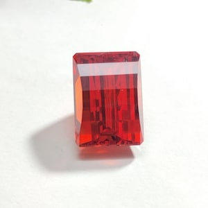 Pode incluir: Uma pedra preciosa vermelha, lapidada em forma retangular e facetada. A gema é transparente, com facetas internas que criam um jogo de luz. A pedra preciosa está sobre um fundo branco.