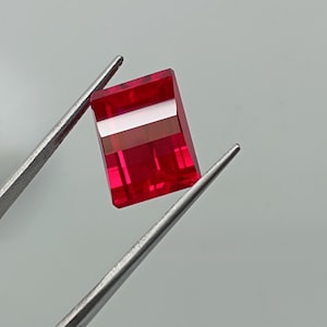 May include: A rectangular, faceted gemstone with a vibrant, deep red colour held by a pair of silver-toned metal tweezers. The stone features sharp, clean edges and a polished surface that reflects light, showcasing its transparent and saturated red hue.