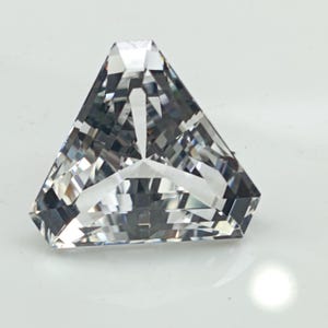 Può includere: Una gemma trasparente e sfaccettata a forma triangolare. La gemma ha molteplici sfaccettature che riflettono la luce, creando un effetto scintillante. La gemma è posta su uno sfondo bianco.