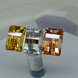 Può includere: Tre pietre preziose rettangolari nelle tonalità del giallo, trasparente e arancione. Le gemme sono sfaccettate, riflettono la luce e creano un effetto scintillante. Le gemme sono esposte su una superficie riflettente.