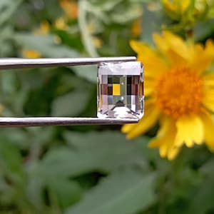 Può includere: Una gemma trasparente di forma rettangolare con taglio a smeraldo, tenuta da una pinzetta in metallo color argento. La pietra presenta precise sfaccettature geometriche che riflettono la luce, creando un aspetto dai toni multipli.