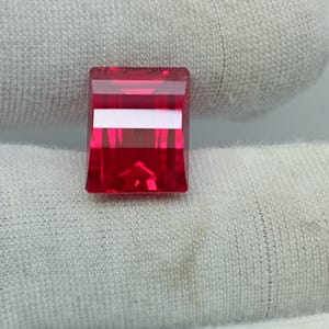 Peut inclure: Une pierre précieuse rectangulaire d'un rouge profond avec des surfaces facettées créant des reflets lumineux nets. La pierre présente un aspect transparent et clair avec un reflet lumineux blanc horizontal distinct au centre.