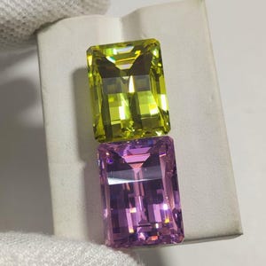 Può includere: Vengono presentate due pietre preziose rettangolari, una verde chartreuse e una lavanda. Le gemme hanno un taglio sfaccettato, che riflette la luce. La gemma verde chartreuse è posizionata sopra la gemma lavanda. Lo sfondo è una superficie bianca.