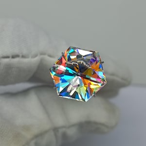 Pode incluir: Uma pedra preciosa hexagonal facetada que exibe um espectro vibrante e iridescente de cores do arco-íris. A pedra apresenta cortes geométricos nítidos que refletem a luz em várias direções, criando um efeito prismático na superfície.