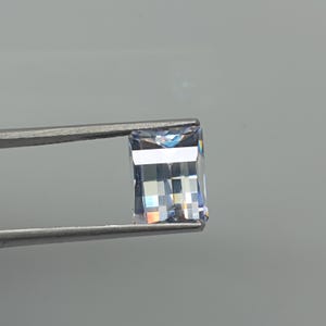Può includere: Una pietra preziosa rettangolare a taglio a gradini, dall'aspetto trasparente e incolore e di elevata purezza. La pietra presenta una rifrazione interna della luce con sottili riflessi arcobaleno sulle faccette, tenuta da una pinzetta metallica.