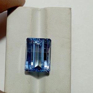 Pode incluir: Uma pedra preciosa retangular e facetada com um tom azul claro. A gema tem uma série de cortes verticais que criam um efeito listrado. A pedra preciosa está sobre um fundo cinza claro.