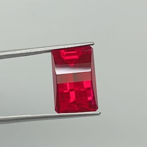 Op de afbeelding: Een levendige, rechthoekig geslepen rode edelsteen vastgehouden door een zilverkleurige metalen pincet. De steen heeft een gefacetteerd oppervlak dat licht reflecteert en een diepe, verzadigde karmozijnrode kleur toont in de kristalstructuur.