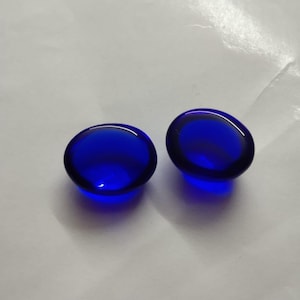 May include: Two oval-shaped, deep blue glass stones. The stones have a smooth, glossy surface and are positioned side by side. The color is a rich, saturated blue, and the stones appear to be translucent. The background is a light, neutral color.
