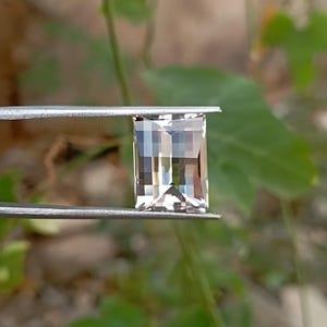 以下が含まれることがあります： 淡い半透明のピンクベージュの色合いをした、長方形のステップカットの宝石。石の表面はファセットカットされており、光を反射して、その透明な結晶構造全体に光と影の幾何学的なパターンを作り出しています。