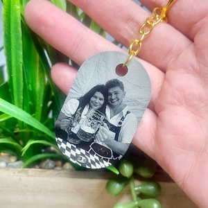 May include: Oval keyring with a gold-coloured ring and chain. The keyring displays a black and white image of a couple holding beer steins. The background is blurred, with green plants visible.