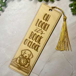 May include: A wooden bookmark with the engraved text "OH LOOK! it's BOOK O'CLOCK". The design features a dog with a clock on an open book. A gold tassel is attached to the top of the bookmark.