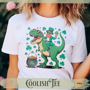 Könnte beinhalten: Weißes T-Shirt mit einem grünen Dinosaurier mit Sonnenbrille und einem Leprechaun, der ihn reitet. Der Leprechaun hält ein Kleeblatt und trägt einen grünen Anzug und Hut. Ein Topf mit Gold und Kleeblättern sind ebenfalls im Design. Der Markenname "Coolish Tee" steht unten.