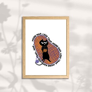 May include: A framed art print featuring a black cat holding yarn inside an orange and purple net. The text "MORE NAP, LESS TRAP" is written around the net. The artwork has a minimalist, cartoon style.