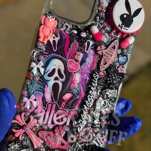May include: A clear phone case decorated with a variety of embellishments. The design includes a Scream mask, pink accents, and the word "Killer". Additional elements include a Playboy bunny logo and various charms.