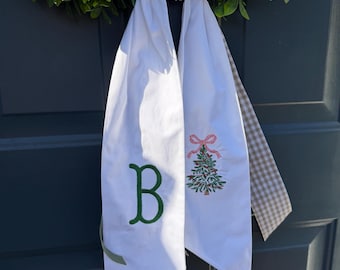 May include: White fabric door hanger with a green embroidered letter "B" and a Christmas tree design. The hanger is tied to a green wreath, creating a festive holiday decoration.