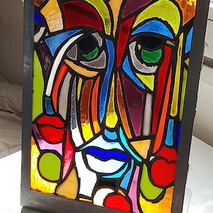 Puede incluir: Obra de arte abstracta en vidriera que representa un rostro estilizado con líneas audaces y colores vibrantes. La obra presenta una variedad de colores, incluyendo rojo, azul, verde, amarillo y morado, enmarcada en un marco gris oscuro. La pieza se exhibe sobre una superficie blanca.