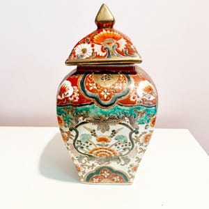 May include: A decorative ceramic jar with a lid. The jar is decorated with a floral pattern in shades of red, green, and gold. The lid has a gold finial.
