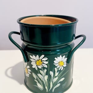 May include: A green metal planter with two handles, painted with white daisies and yellow centers. The planter has a brown terracotta inner pot.