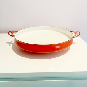 May include: A red enamel cast iron baking dish with two handles. The dish has a white interior.