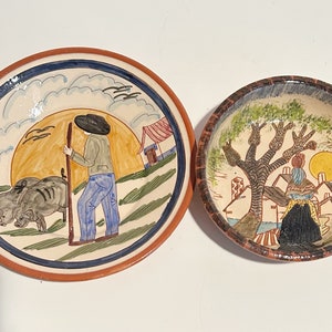 Peut inclure: Deux assiettes en céramique avec des scènes peintes à la main. Une assiette représente un homme avec un bâton marchant avec deux cochons dans un champ. L'autre assiette représente une femme debout sous un arbre avec des maisons en arrière-plan.