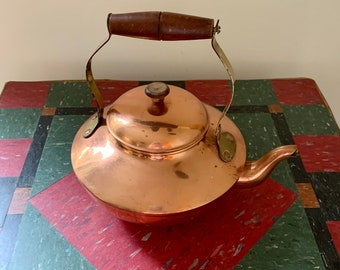 May include: A copper tea kettle with a wooden handle and brass accents. The kettle is sitting on a red and green checkered surface.