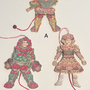 Op de afbeelding: Drie kleurrijke houten trekpoppen. De eerste pop is een persoon die een kleurrijk kostuum draagt met een zon op zijn hoofd. De tweede pop is een persoon die een groen en roze kostuum draagt met appels en manden. De derde pop is een persoon die een roze en bruin kostuum draagt met donuts.