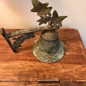 May include: A wall-mounted bell with a weathered bronze finish, featuring a decorative bracket adorned with sculpted ivy leaves. The bell itself has a flared rim and a matching leaf-shaped finial at the top, displaying a dark, aged patina.