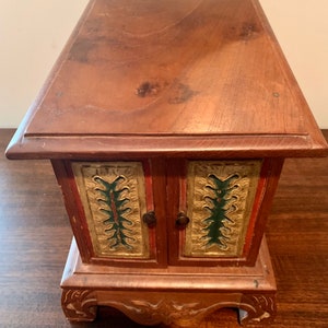 May include: A small wooden box with two doors, each decorated with gold and green painted designs. The box has a carved base and sits on a wooden surface.