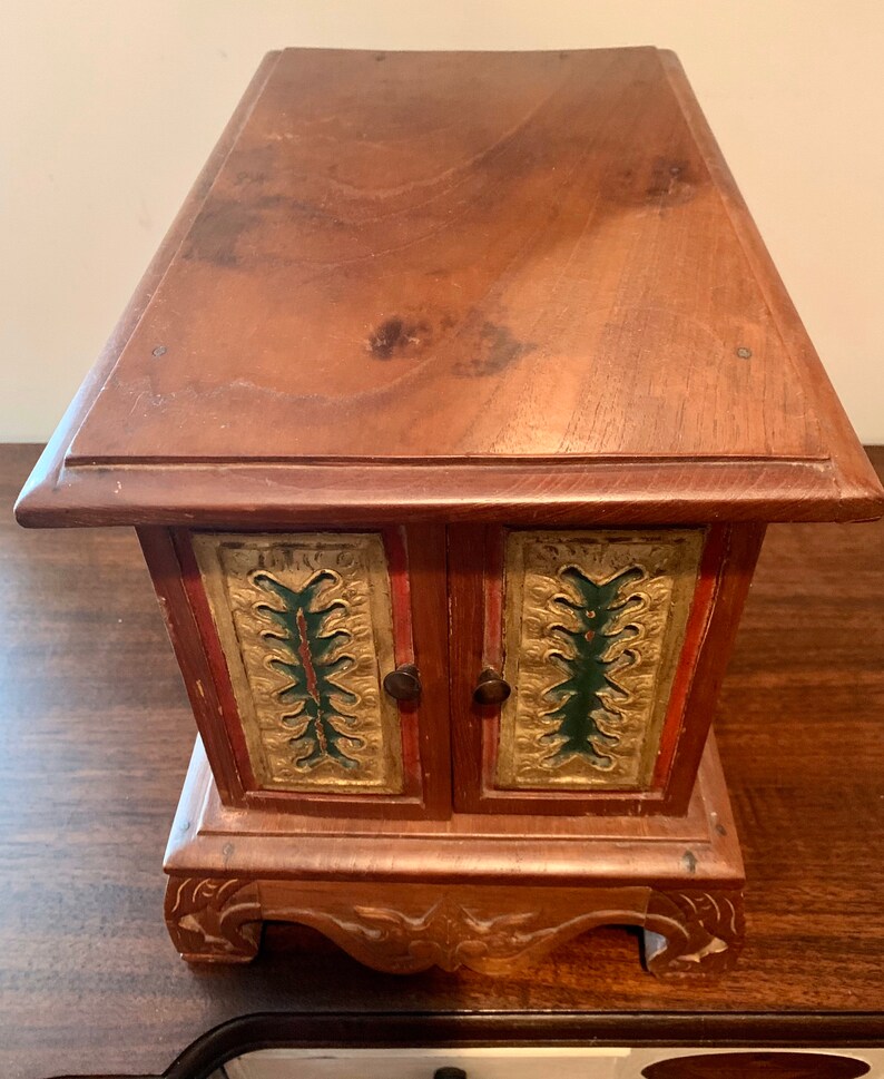 May include: A small wooden box with two doors, each decorated with gold and green painted designs. The box has a carved base and sits on a wooden surface.