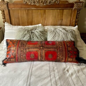 May include: A rectangular decorative pillow with a red and brown geometric pattern, accented with black fringe on the ends. The pillow is placed on a bed with a wooden headboard and fluffy gray pillows.