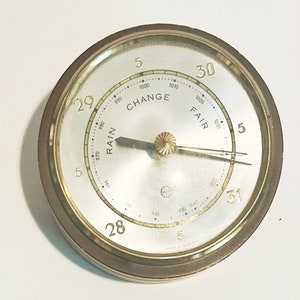 May include: A round, brass-colored barometer with a white face and black markings. The face has a needle that points to the current weather conditions, which are indicated by the words "RAIN", "CHANGE", and "FAIR". The numbers 28, 29, 30, and 31 are printed around the edge of the face.