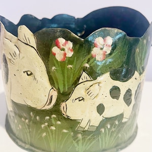 Op de afbeelding: Een handbeschilderde keramische plantenbak met een groene achtergrond en een wit varken en een kleiner wit varken met zwarte vlekken. De varkens zijn omringd door groen gras en roze bloemen.